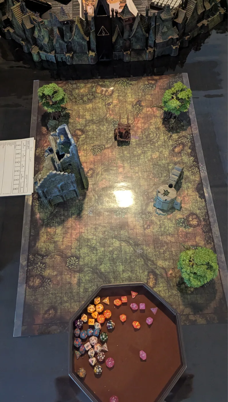 A battle map set up with terrain, ruins and a tray of dice